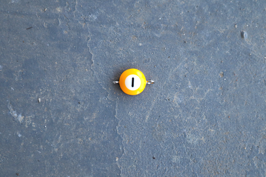 A yellow pool ball pendant with silver rings attached to the right and left side of the pendant. A "1" is in the center of the pendant which is resting on a blue surface.