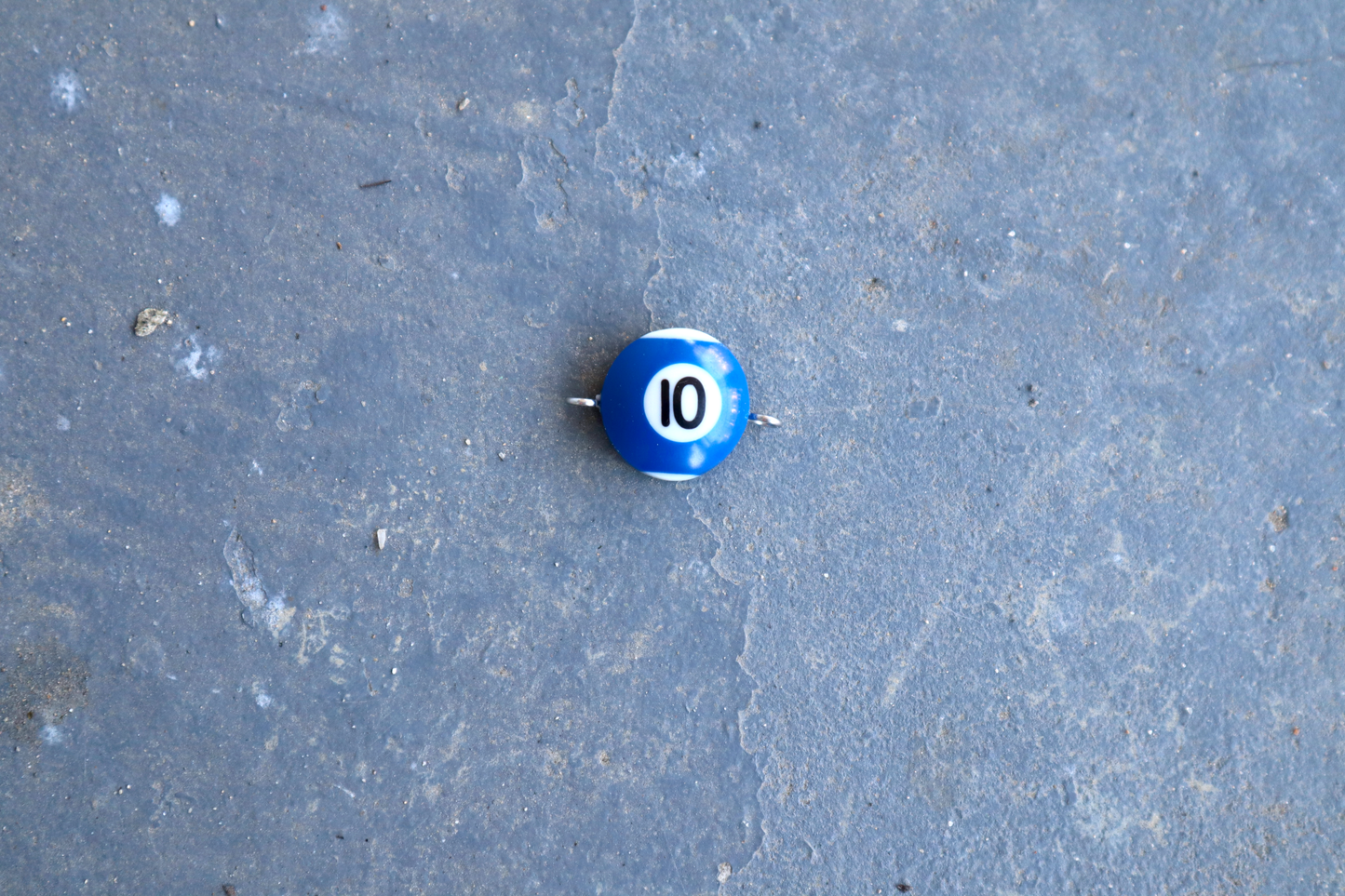 A blue pool ball pendant with silver rings attached to the right and left side of the pendant. A "10" is in the center of the pendant which is resting on a blue surface.