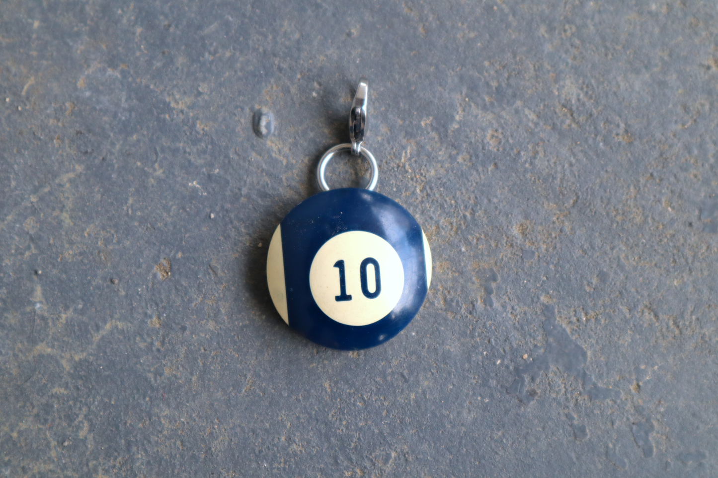 a blue and white billiard ball pendant marked "10" in the center. A silver clasp is attached to the top of the pendant against a blue/gray surface