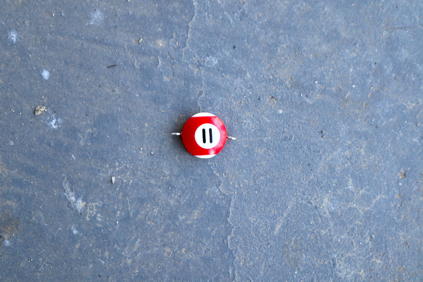 A red and white pool ball pendant with silver rings attached to the right and left side of the pendant. A "11" is in the center of the pendant which is resting on a blue surface.