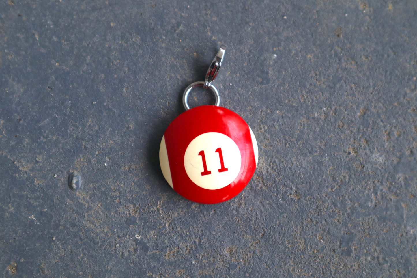 a red and white billiard ball pendant marked "11" in the center. A silver clasp is attached to the top of the pendant against a blue/gray surface