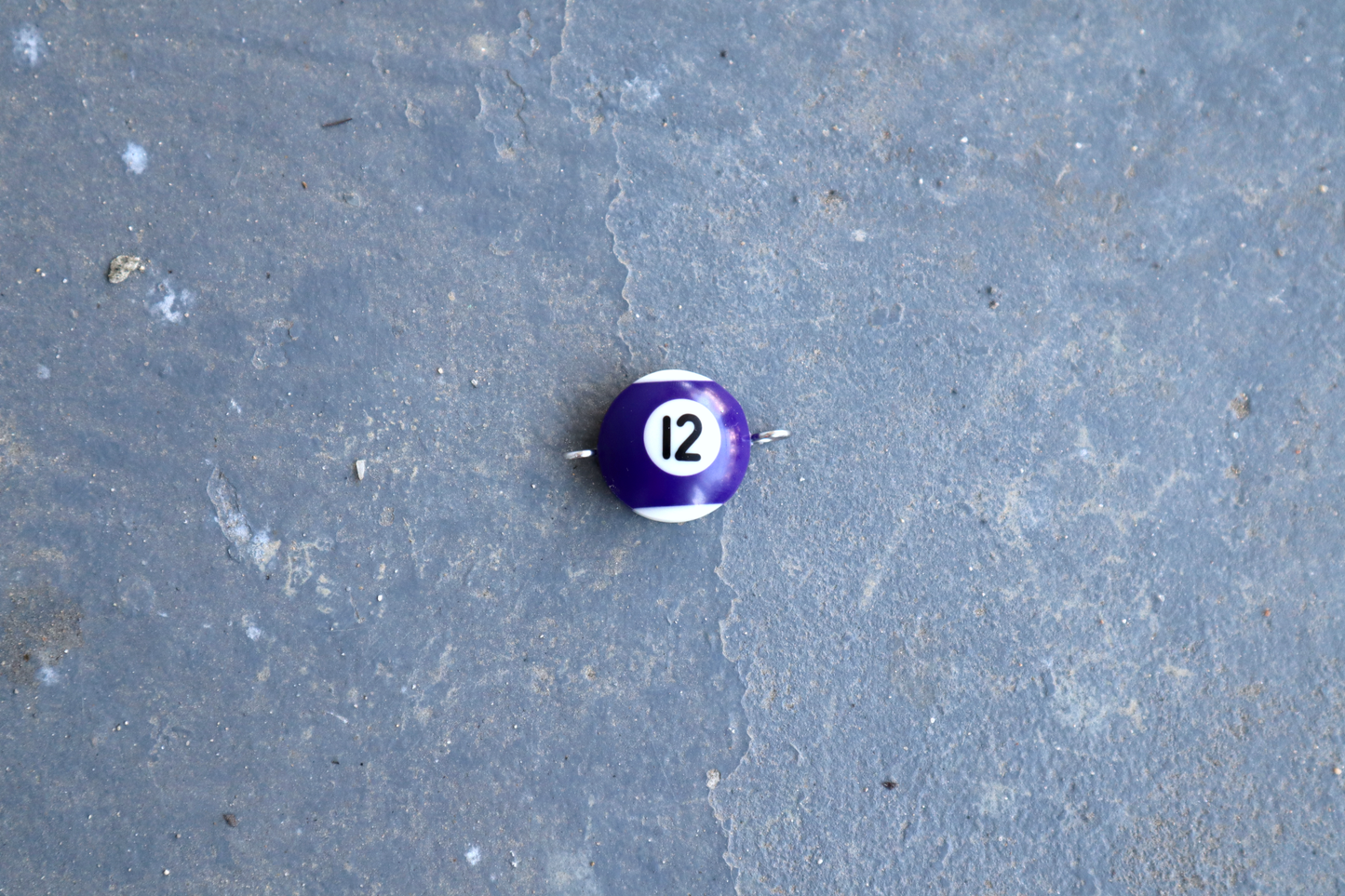 A purple and white pool ball pendant with silver rings attached to the right and left side of the pendant. A "12" is in the center of the pendant which is resting on a blue surface.