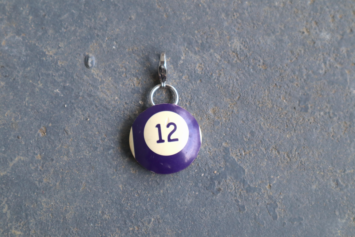 a purple and white billiard ball pendant marked "12" in the center. A silver clasp is attached to the top of the pendant against a blue/gray surface
