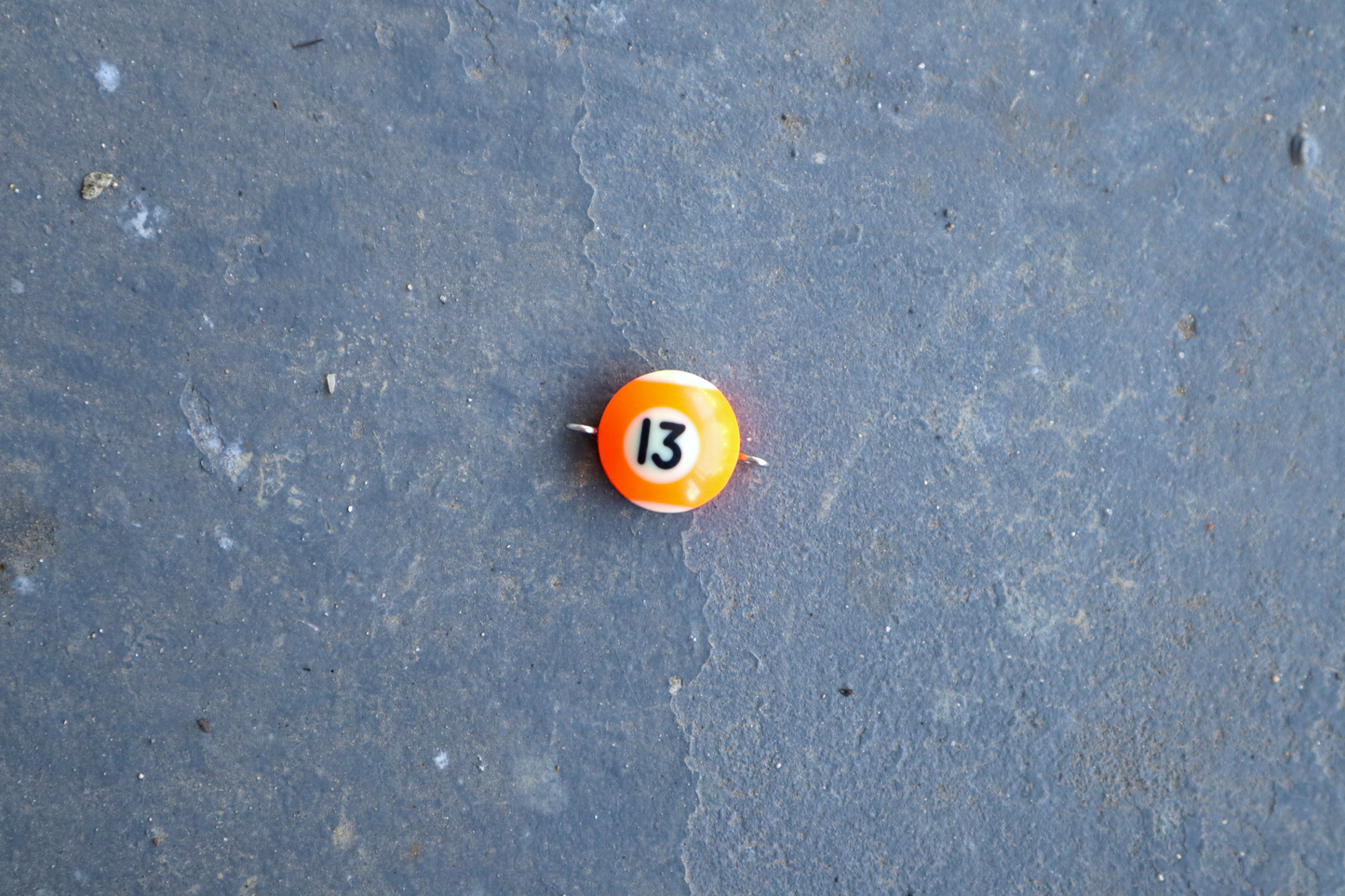 An orange and white pool ball pendant with silver rings attached to the right and left side of the pendant. A "13" is in the center of the pendant which is resting on a blue surface.