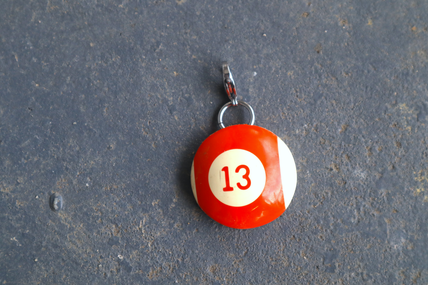 an orange and white billiard ball pendant marked "13" in the center. A silver clasp is attached to the top of the pendant against a blue/gray surface