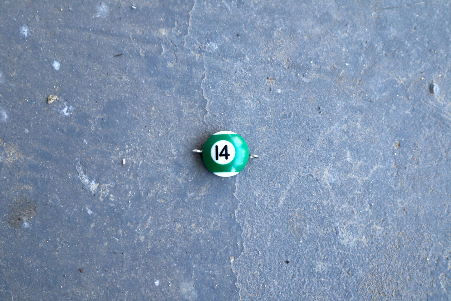 A green and white pool ball pendant with silver rings attached to the right and left side of the pendant. A "14" is in the center of the pendant which is resting on a blue surface.