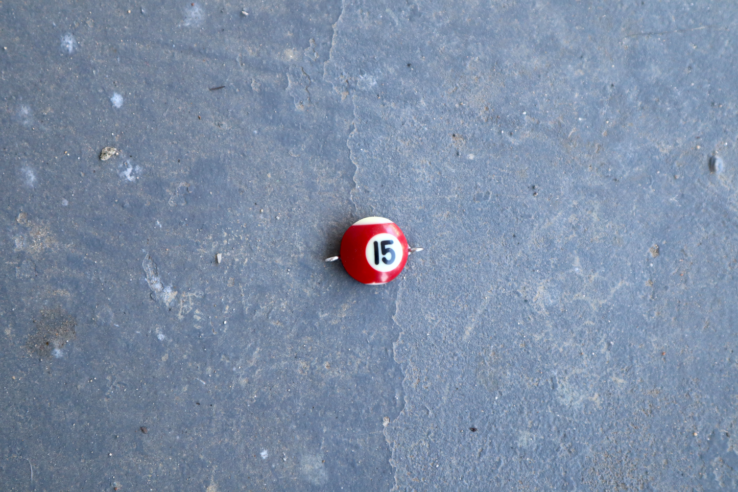 A red and white pool ball pendant with silver rings attached to the right and left side of the pendant. A "15" is in the center of the pendant which is resting on a blue surface.