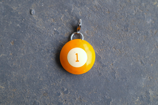 a yellow billiard ball pendant marked "1" in the center. A silver clasp is attached to the top of the pendant against a blue/gray surface
