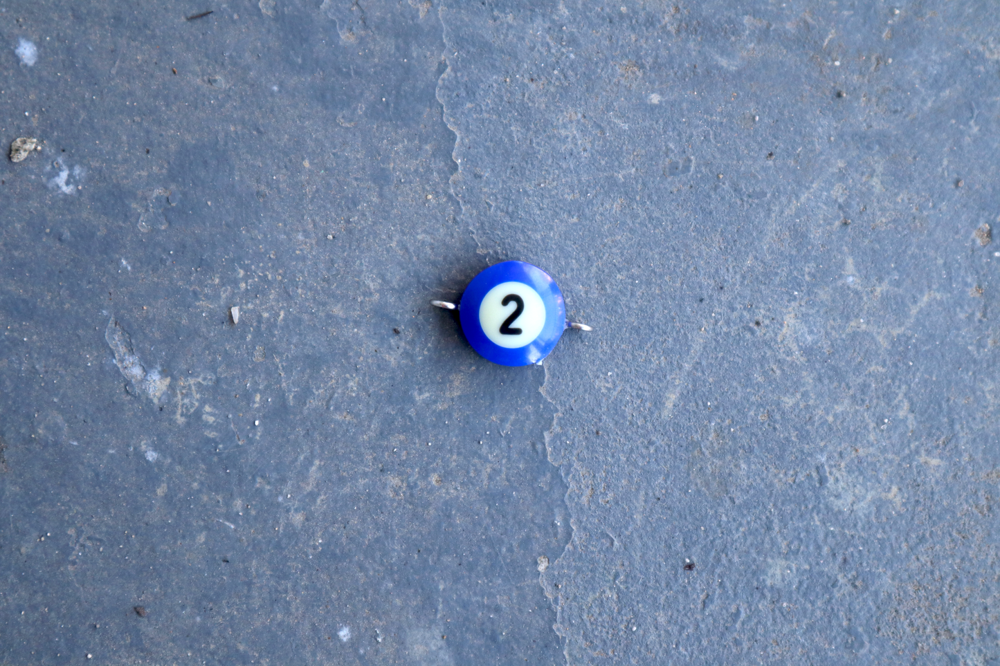 A blue pool ball pendant with silver rings attached to the right and left side of the pendant. A "2" is in the center of the pendant which is resting on a blue surface.