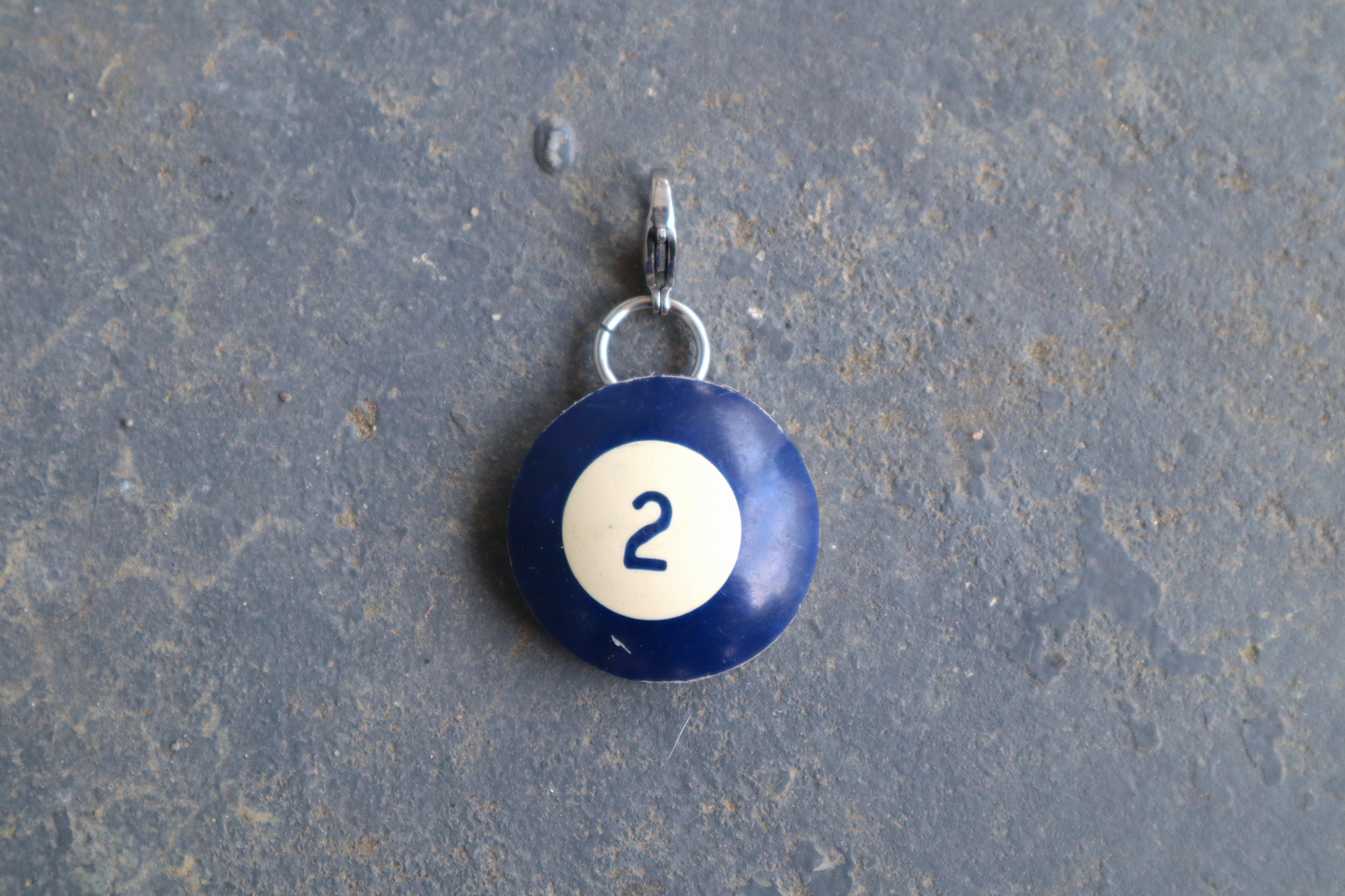 a blue billiard ball pendant marked "2" in the center. A silver clasp is attached to the top of the pendant against a blue/gray surface