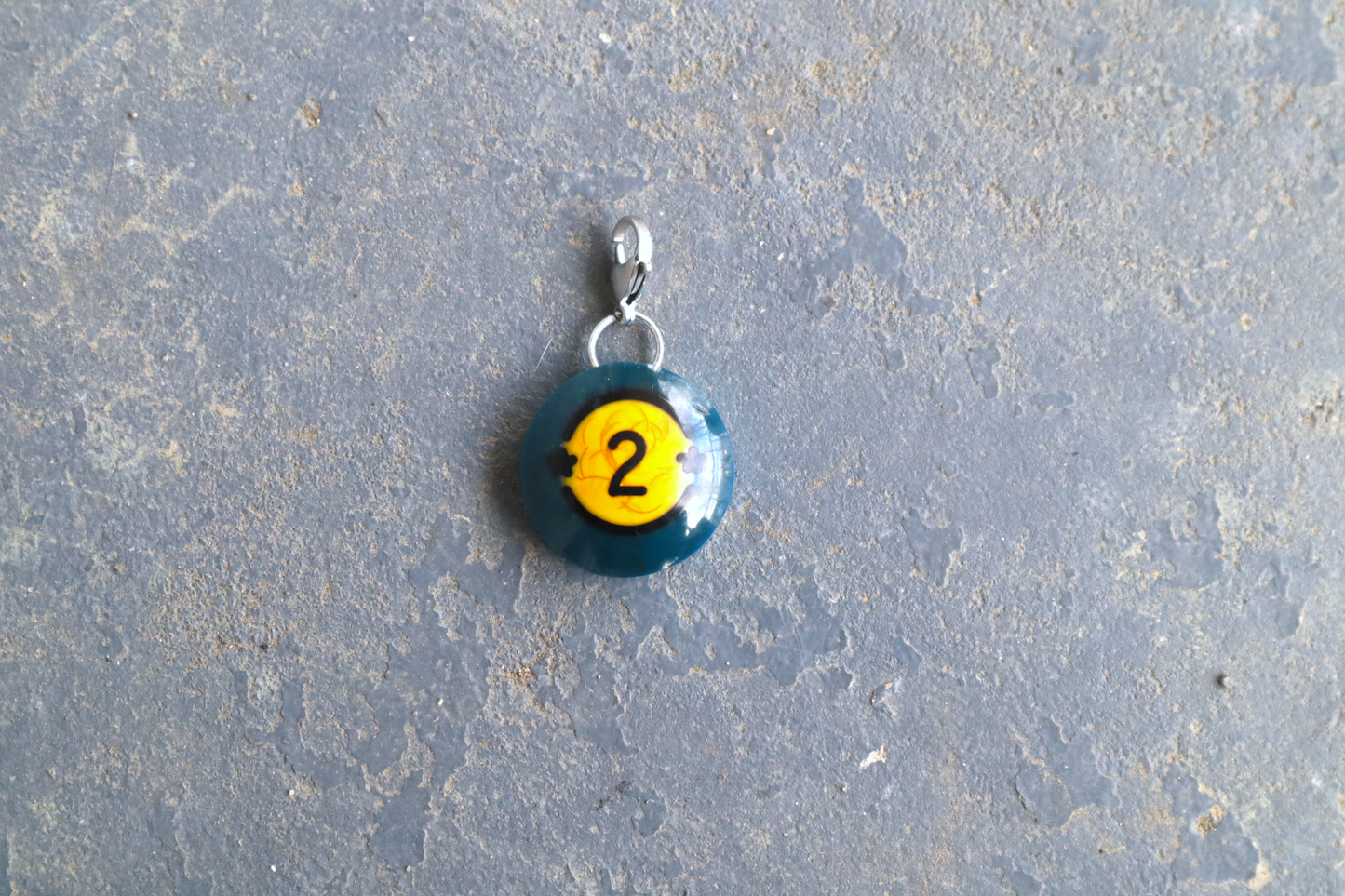 a blue billiard ball pendant marked "2" with 2 black stars on both sides of the number. A silver clasp is attached to the top of the pendant against a blue/gray surface