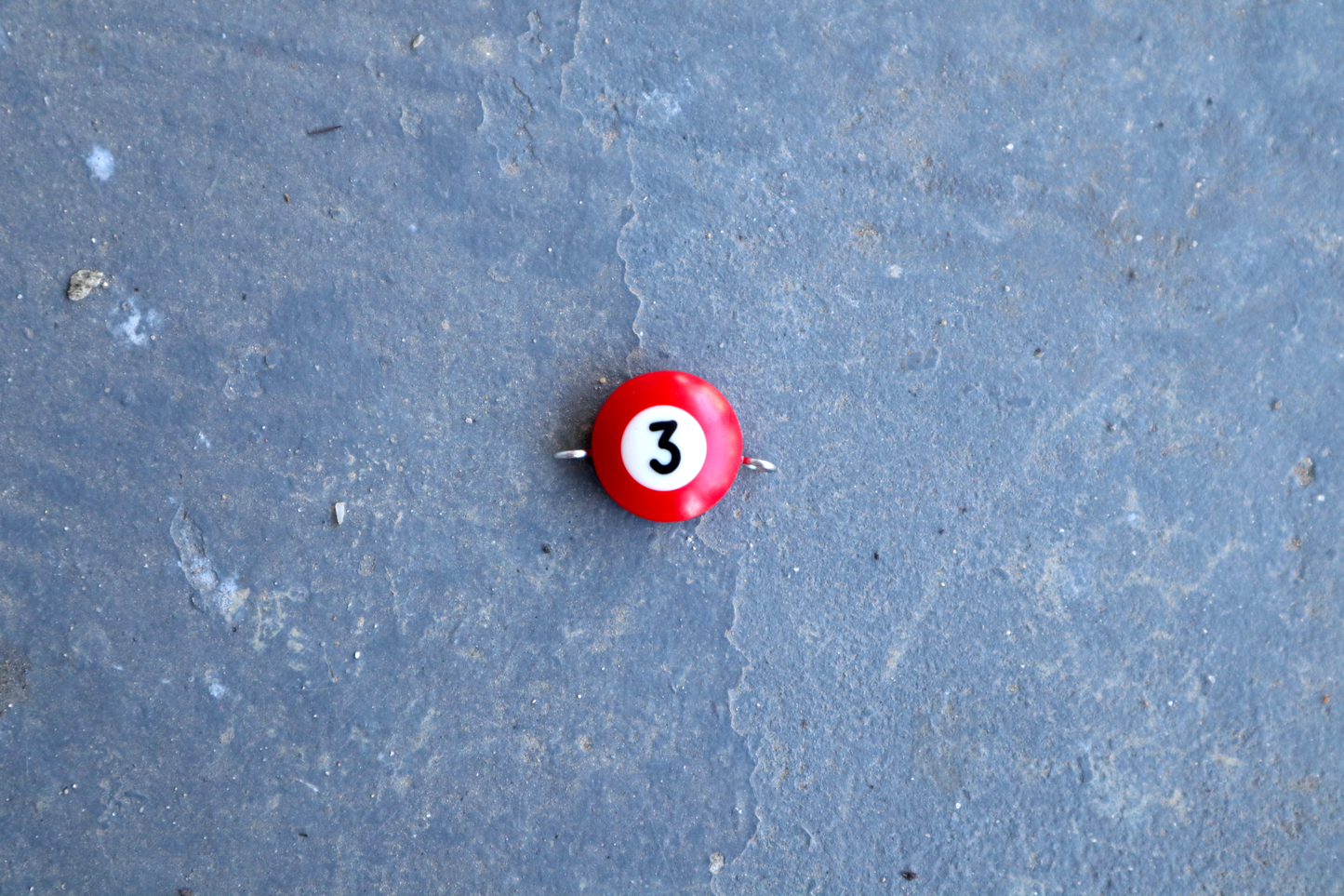 A red pool ball pendant with silver rings attached to the right and left side of the pendant. A "3" is in the center of the pendant which is resting on a blue surface.