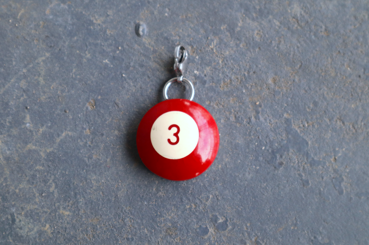 a red billiard ball pendant marked "3" in the center. A silver clasp is attached to the top of the pendant against a blue/gray surface