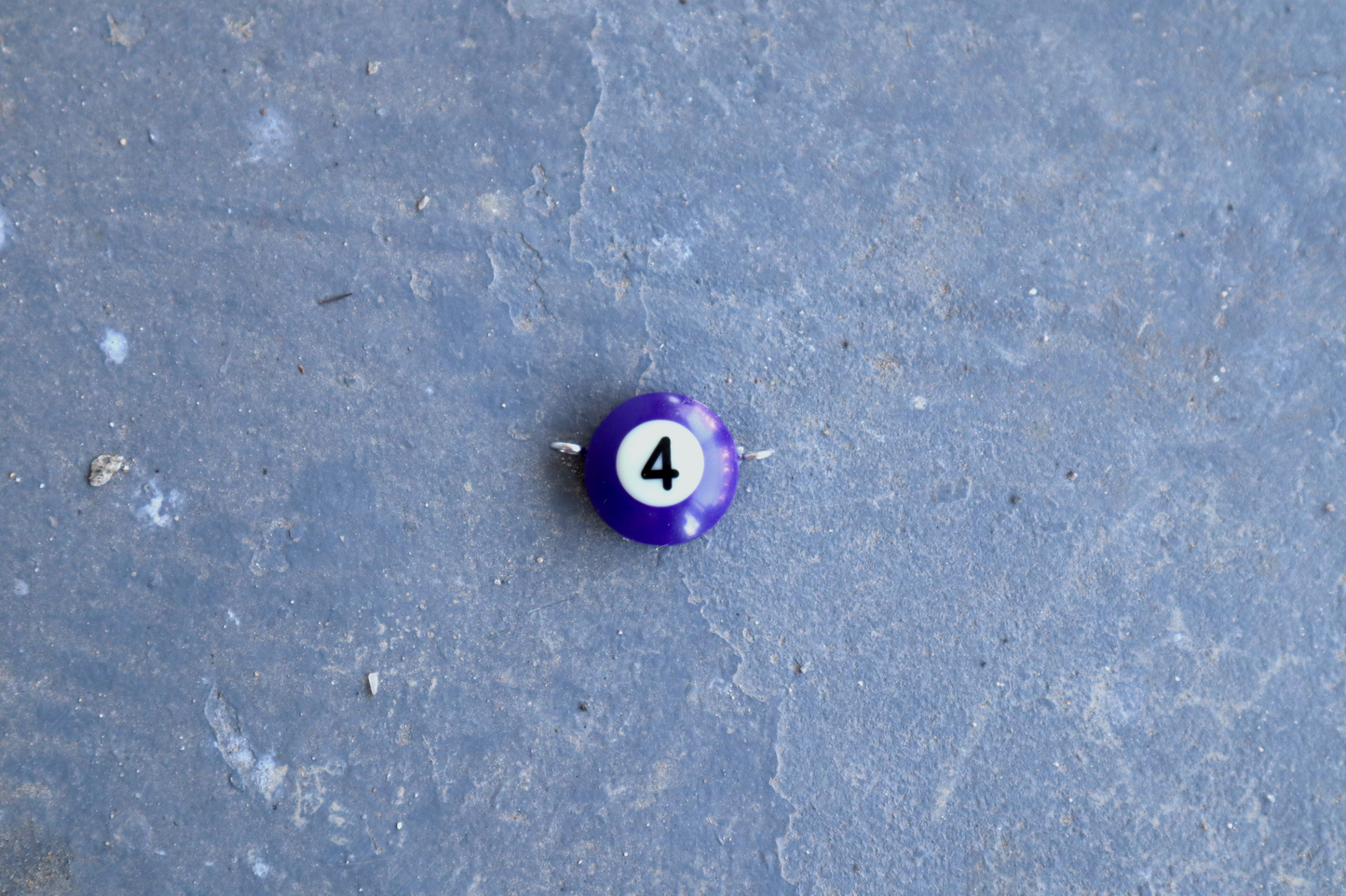 A purple pool ball pendant with silver rings attached to the right and left side of the pendant. A "4" is in the center of the pendant which is resting on a blue surface.