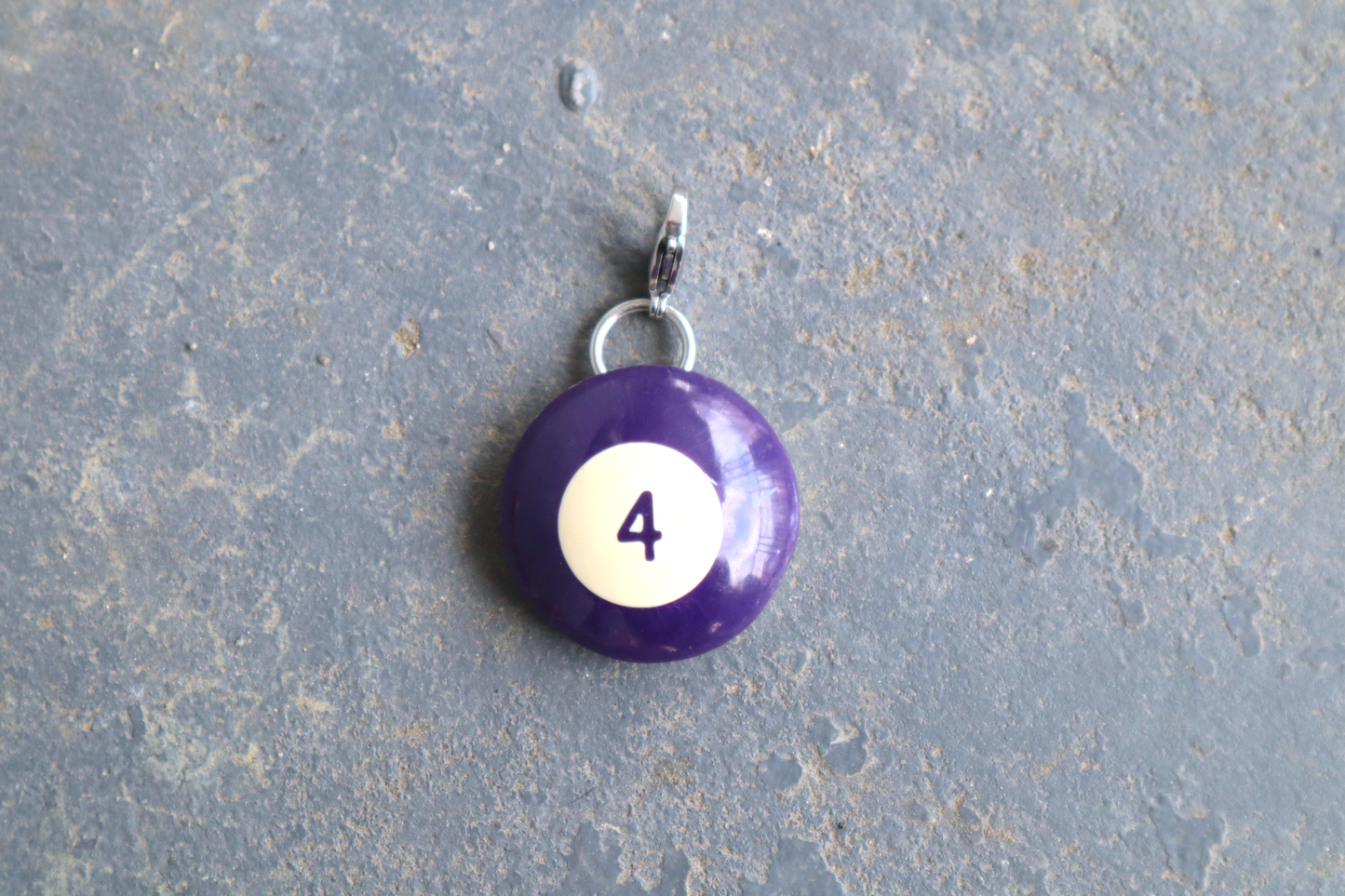 a purple billiard ball pendant marked "4" in the center. A silver clasp is attached to the top of the pendant against a blue/gray surface