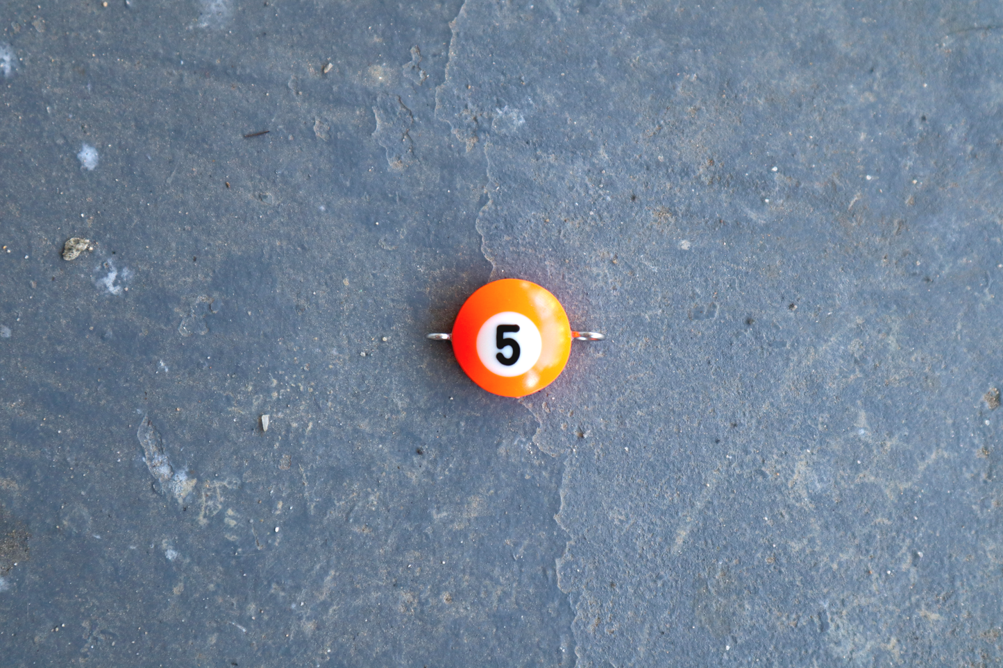 An orange pool ball pendant with silver rings attached to the right and left side of the pendant. A "5" is in the center of the pendant which is resting on a blue surface.