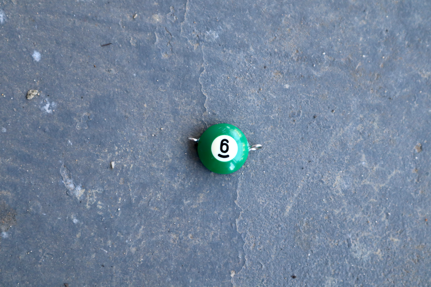 A green pool ball pendant with silver rings attached to the right and left side of the pendant. A "6" is in the center of the pendant which is resting on a blue surface.