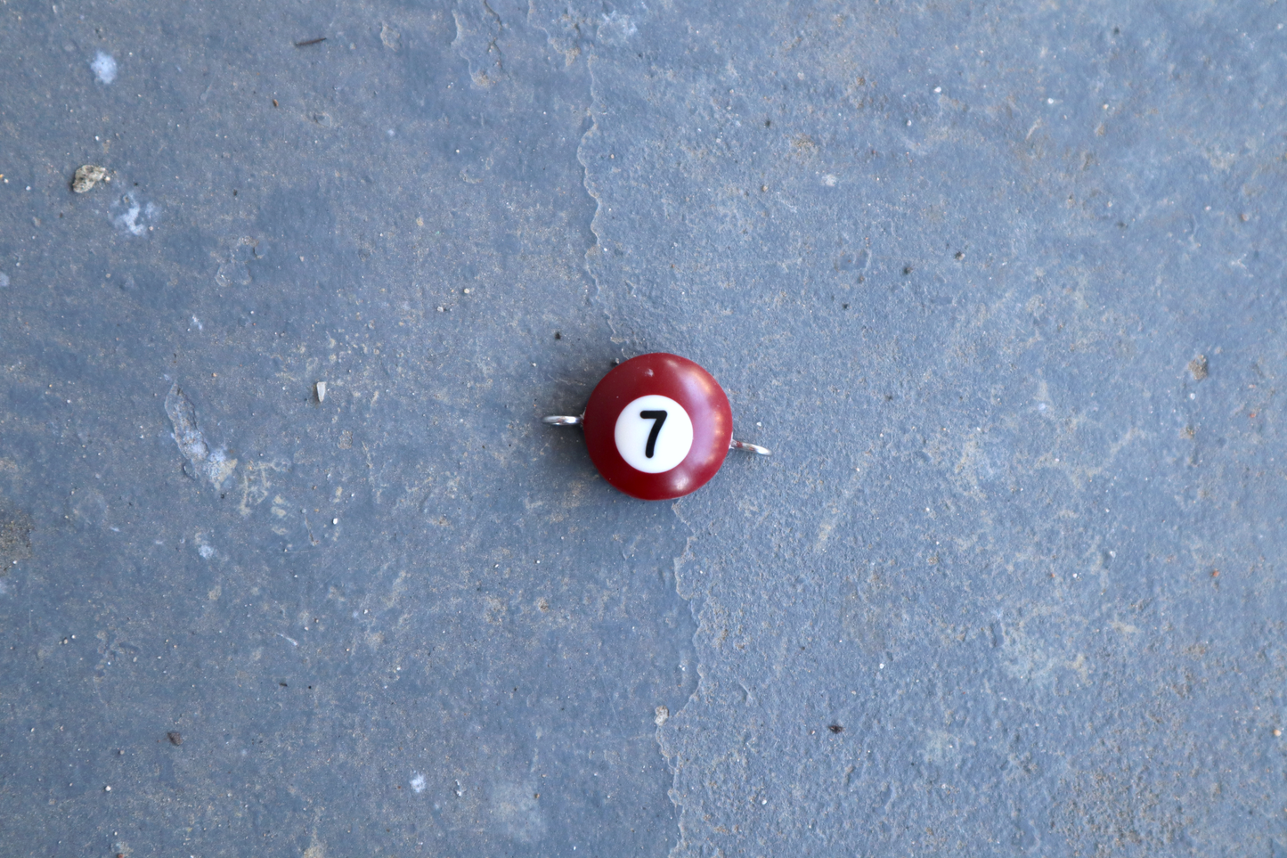 A burgundy pool ball pendant with silver rings attached to the right and left side of the pendant. A "7" is in the center of the pendant which is resting on a blue surface.