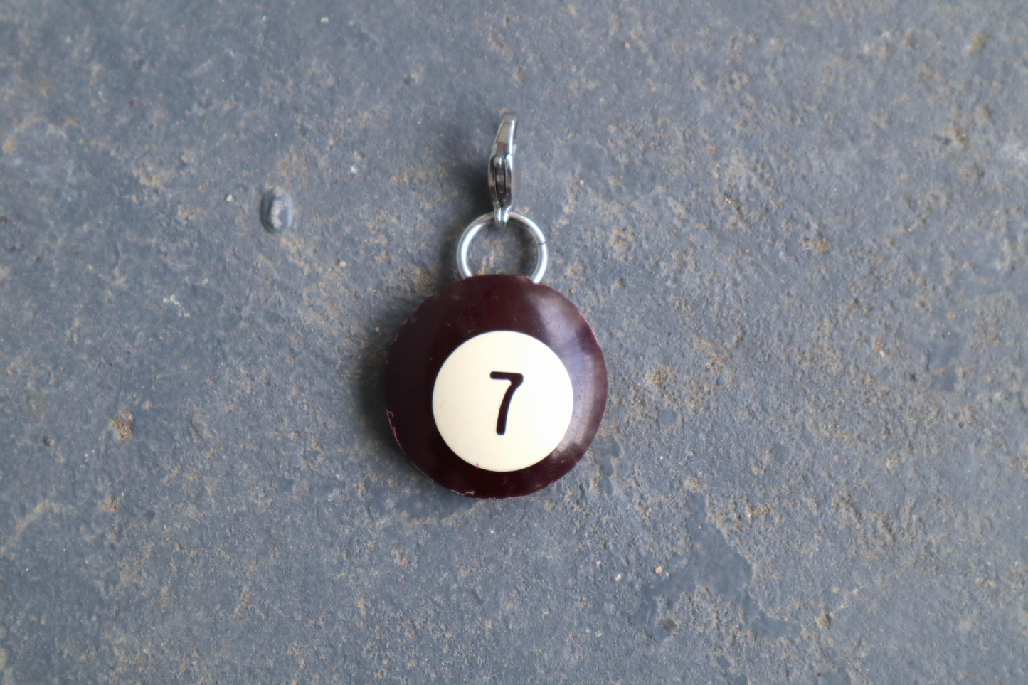 a brown billiard ball pendant marked "7" in the center. A silver clasp is attached to the top of the pendant against a blue/gray surface