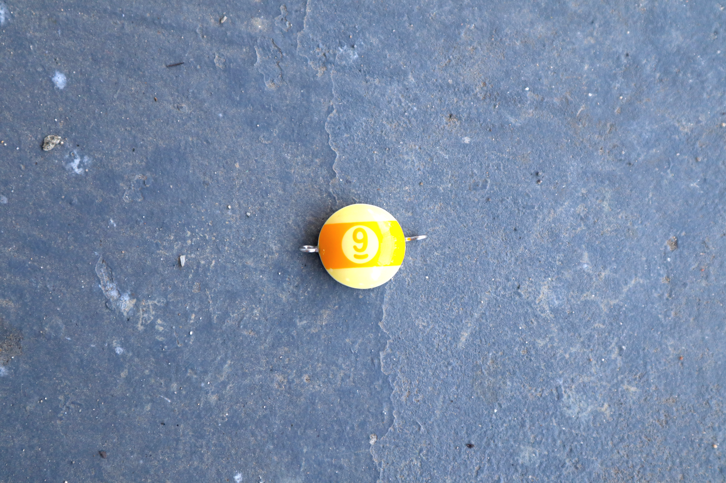 A yellow pool ball pendant with silver rings attached to the right and left side of the pendant. A "9" is in the center of the pendant which is resting on a blue surface.