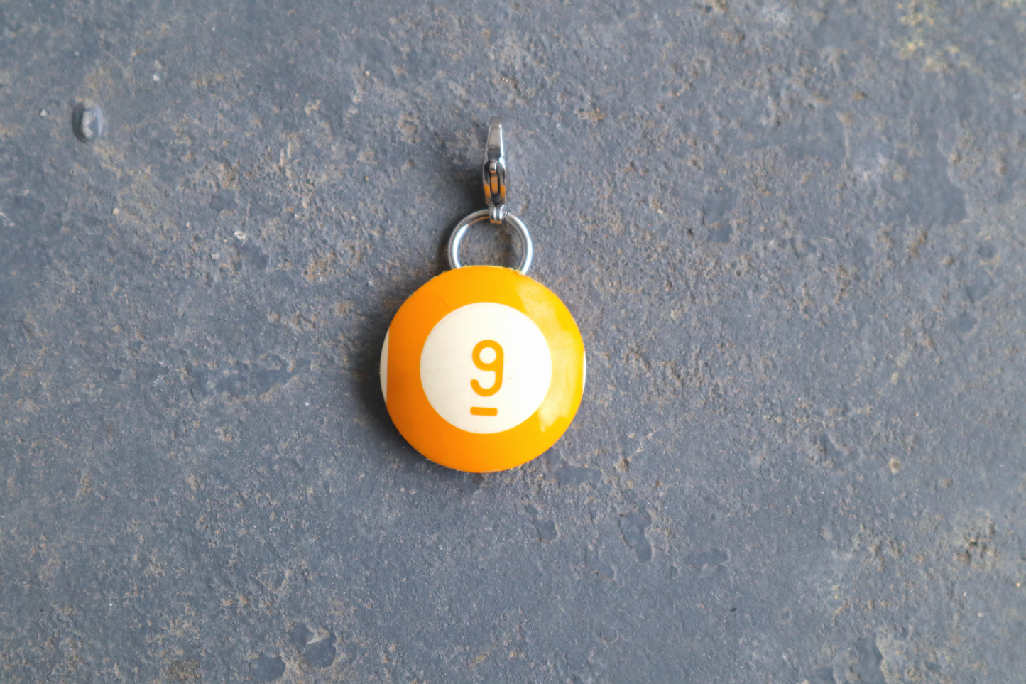 a yellow billiard ball pendant marked "9" in the center. A silver clasp is attached to the top of the pendant against a blue/gray surface