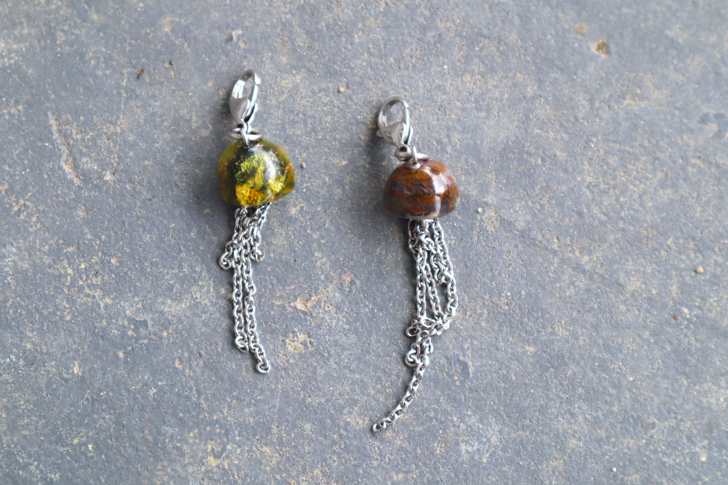 two mushroom cap-shaped stones with a stainless-steel chain hanging from the bottom of them and a steel clasp attached to the tops, against a blue/grey stone background. The stone pendants are orange and green.