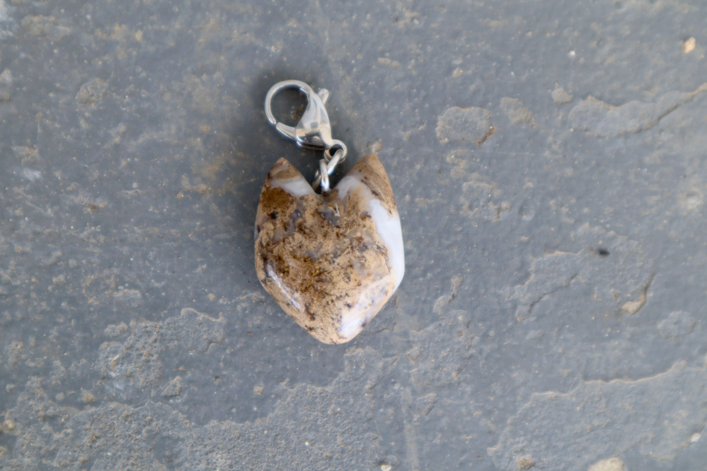 A cat-shaped stone with a stainless-steel clasp attached to it, against a blue/grey stone background. The stone pendant is brown, white and clear in color and speckled with black dots.