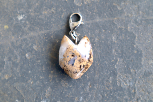 A cat-shaped stone with a stainless-steel clasp attached to it, against a blue/grey stone background. The stone pendant is brown, white and clear in color and speckled with black dots.
