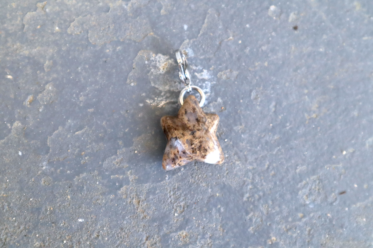 A 5-pointed star-shaped stone with a stainless-steel clasp attached to it, against a blue/grey stone background. The stone pendant is brown, white and clear in color and speckled with black dots.