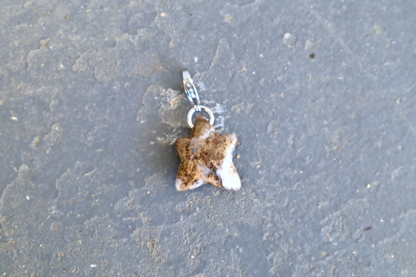 A 5-pointed star-shaped stone with a stainless-steel clasp attached to it, against a blue/grey stone background. The stone pendant is brown, white and clear in color and speckled with black dots.