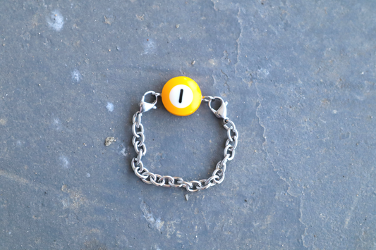 Silver chain bracelet with a yellow billiard ball pendant marked "1" on a blue/gray surface.