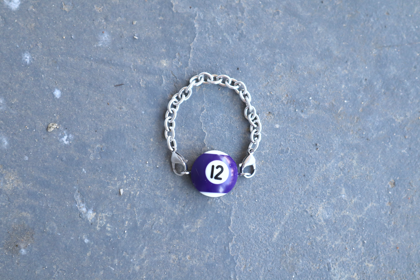 Silver chain bracelet with a purple and white billiard ball pendant marked "12" on a blue/gray surface.