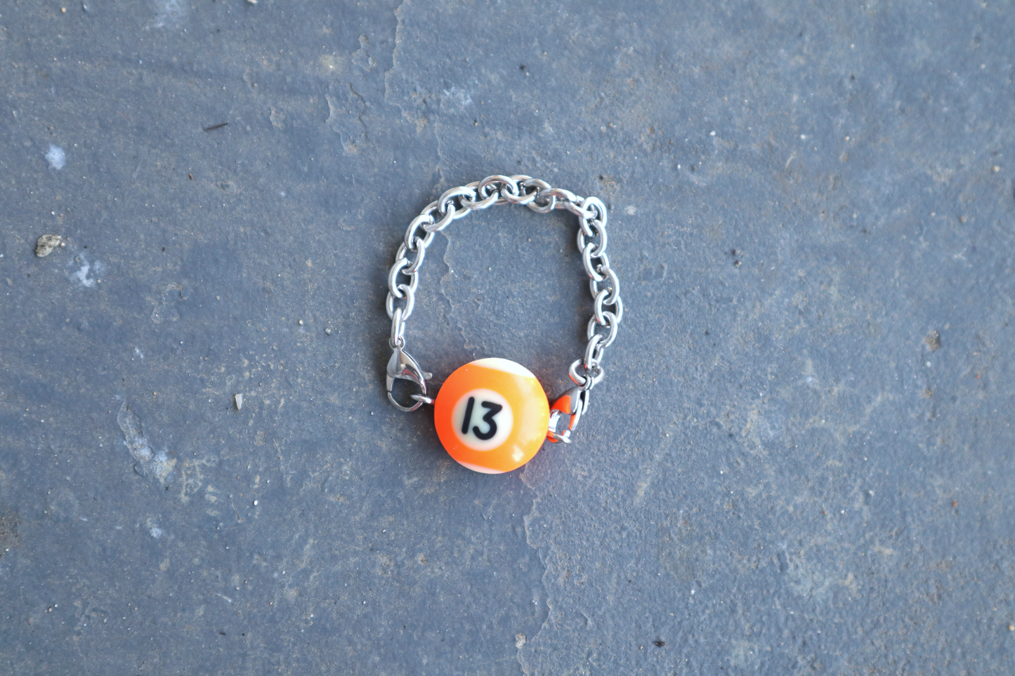 Silver chain bracelet with an orange and white billiard ball pendant marked "13" on a blue/gray surface.
