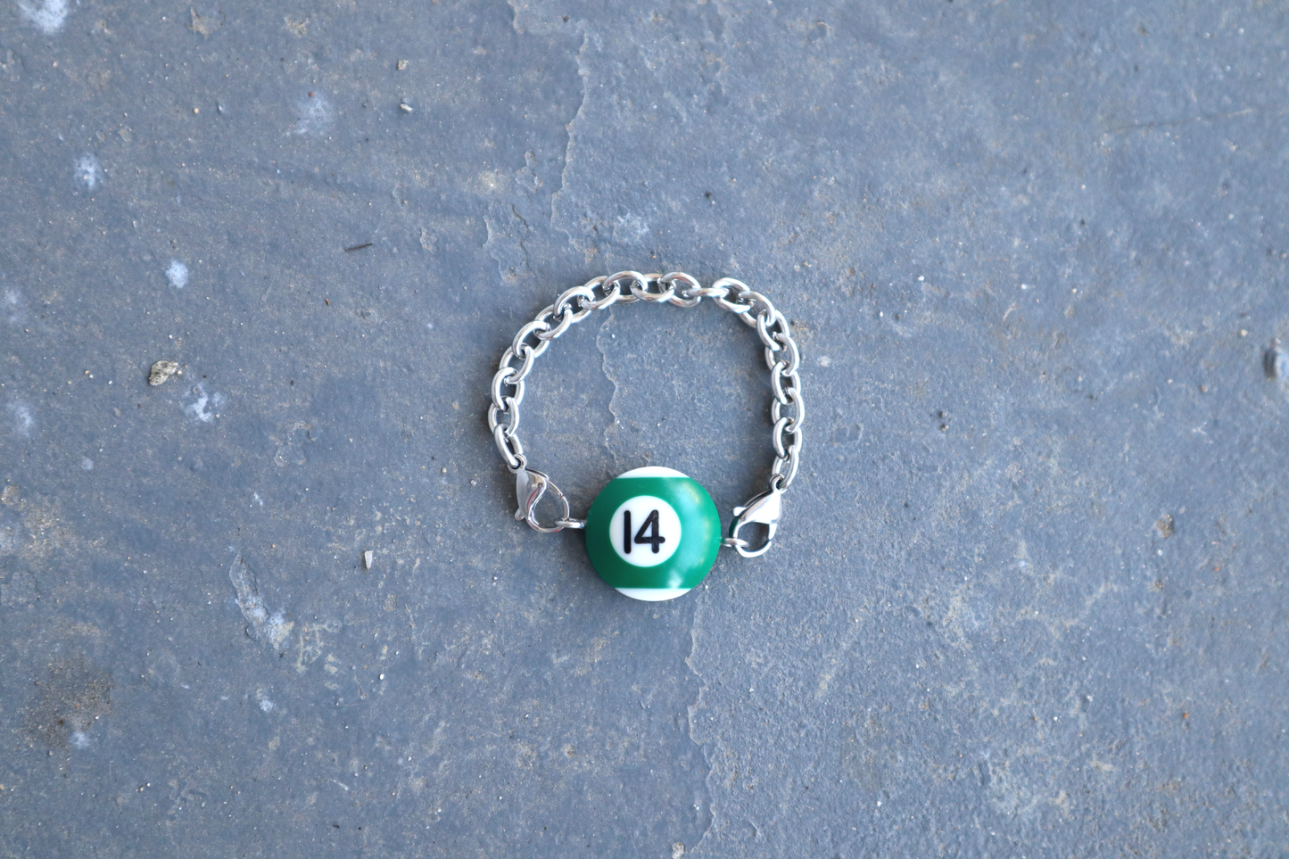 Silver chain bracelet with a green and white billiard ball pendant marked "14" on a blue/gray surface.