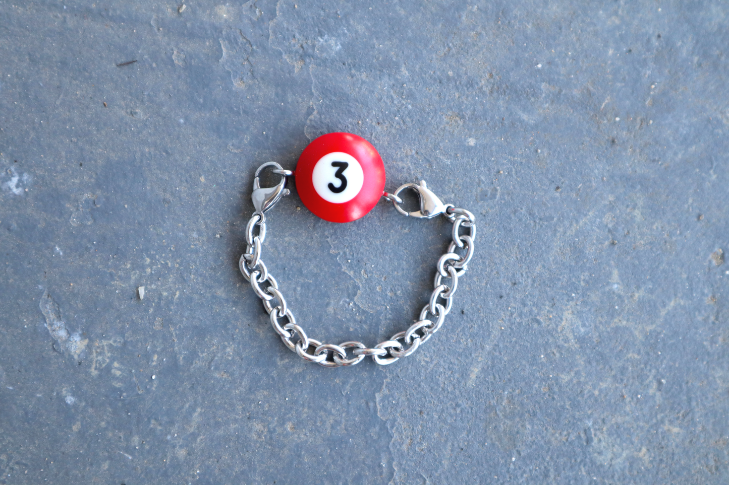 Silver chain bracelet with a red billiard ball pendant marked "3" on a blue/gray surface.