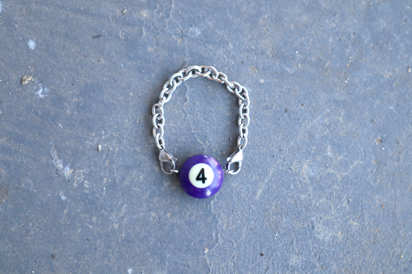 Silver chain bracelet with a purple billiard ball pendant marked "4" on a blue/gray surface.