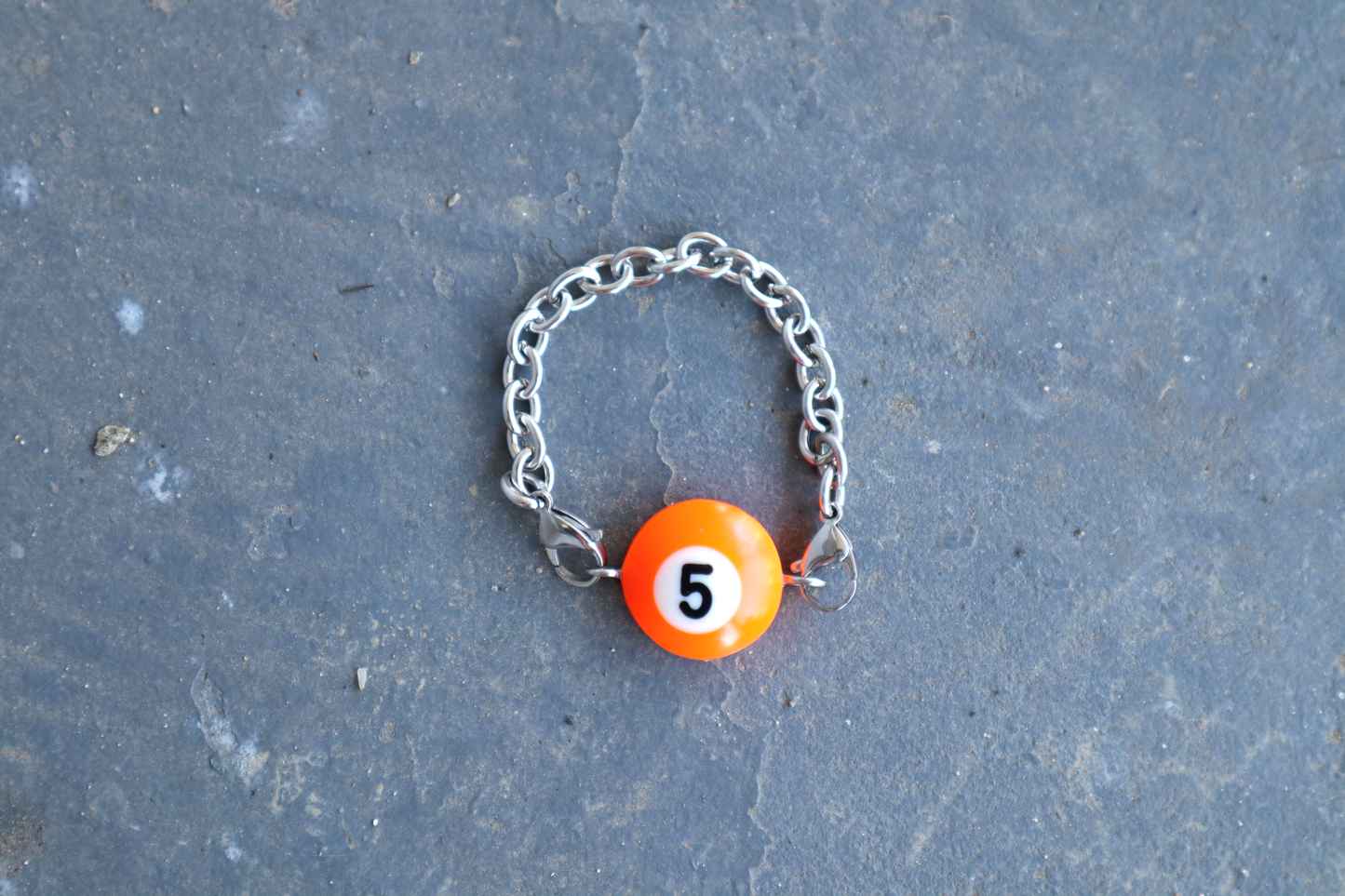Silver chain bracelet with an orange billiard ball pendant marked "5" on a blue/gray surface.