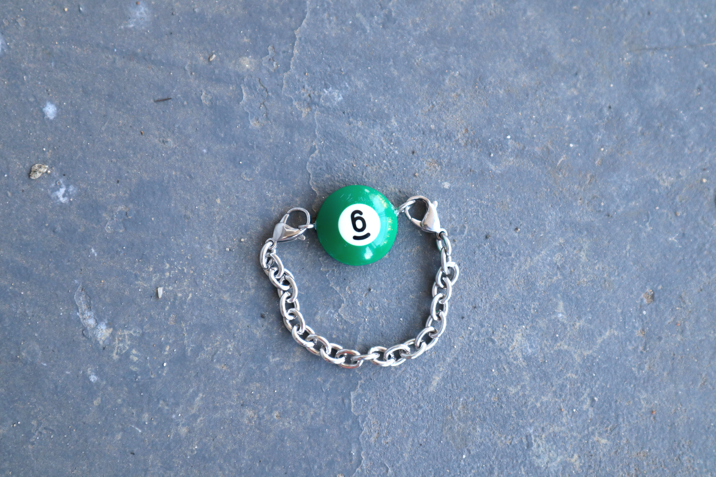 Silver chain bracelet with a green billiard ball pendant marked "6" on a blue/gray surface.