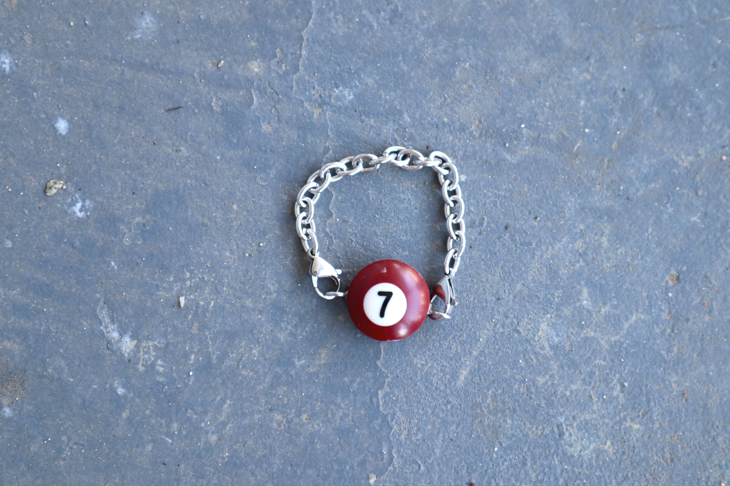 Silver chain bracelet with a burgundy billiard ball pendant marked "7" on a blue/gray surface.