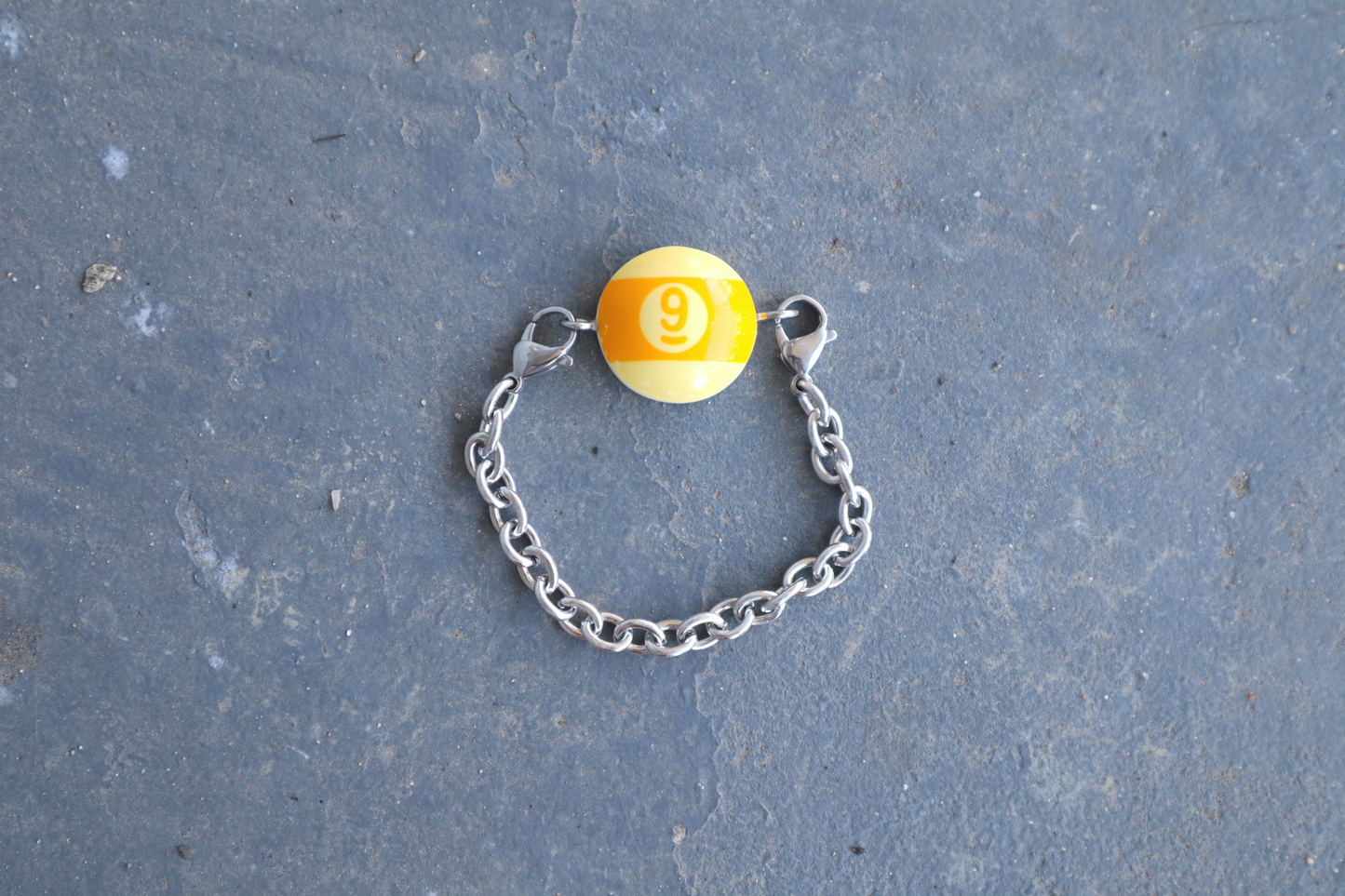 Silver chain bracelet with a yellow and cream billiard ball pendant marked "9" on a blue/gray surface.