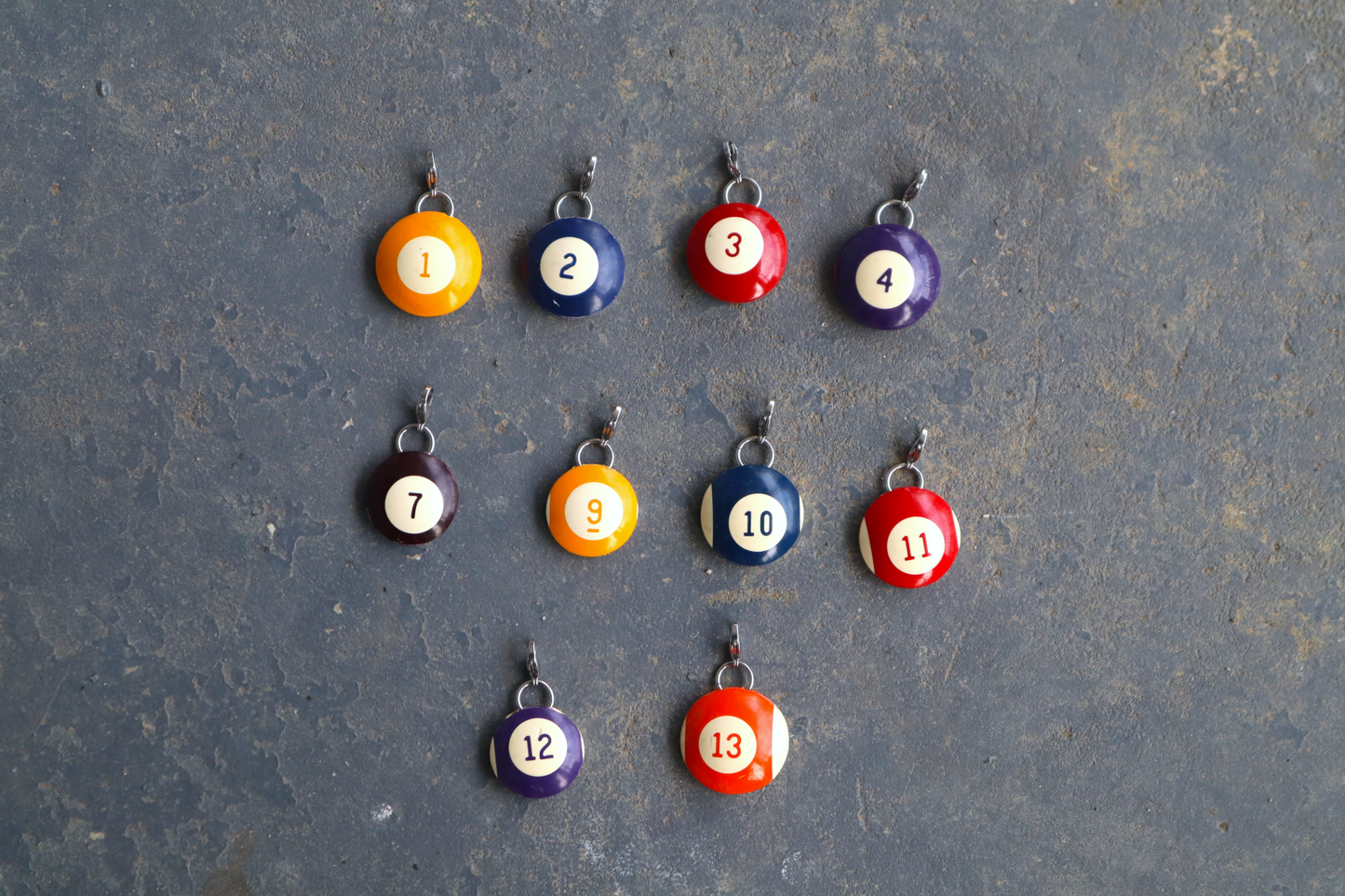 10 billiard ball pendants marked with a number 1 through 15 in the center of each. A silver clasp is attached to the tops of the pendants against a blue/gray surface
