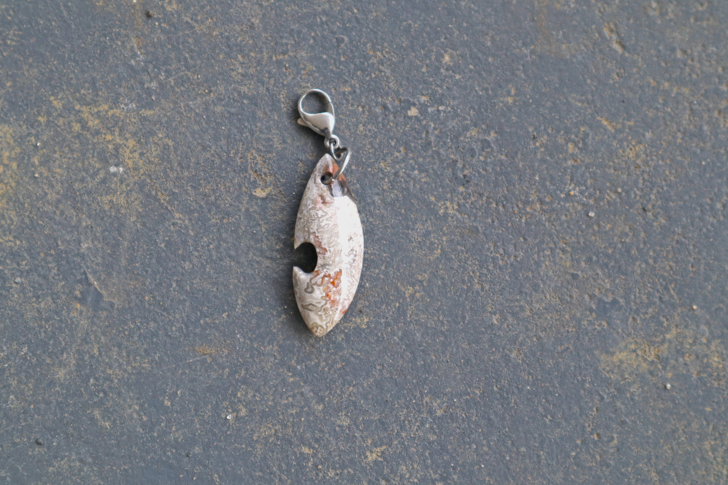 grey, white, black and red a pointed oval-shaped stone with a silver clasp attached to the top of the pendant, resting on a gray surface.