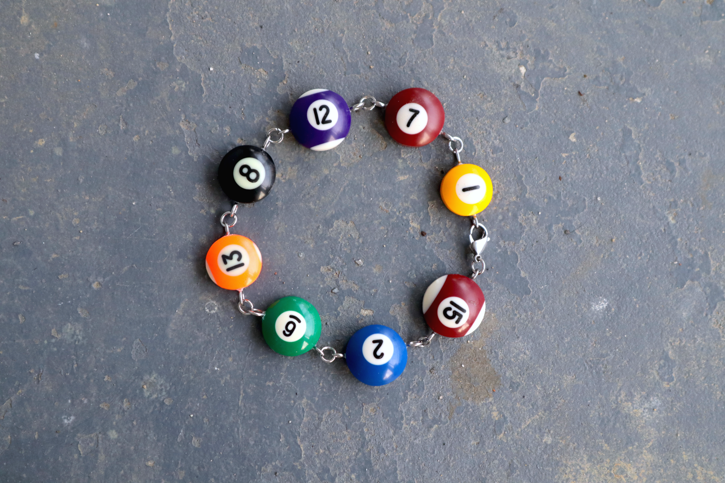 A choker made of pool ball charms numbered 1, 15, 2, 6, 13, 8, 12 and 7 laid against a blue/gray surface. The choker has a steel chain and hardware to connect the charms.