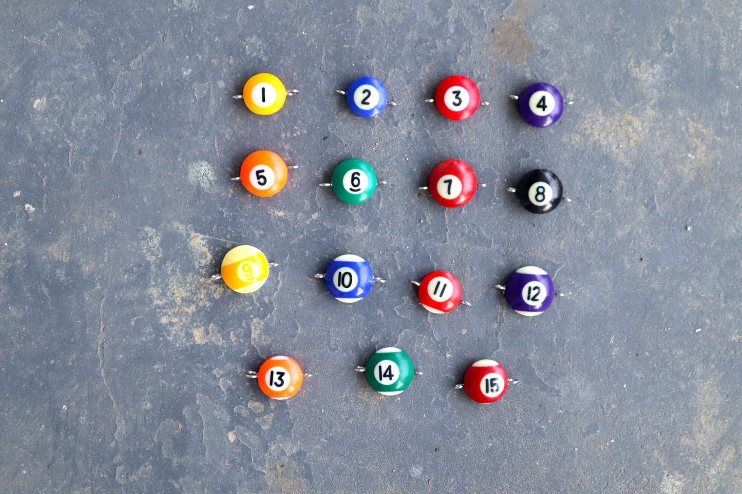 15 pool ball pendants with silver rings attached to the right and left sides of the pendants are spread out across a blue surface. The center of the pendants is numbered 1 through 15.