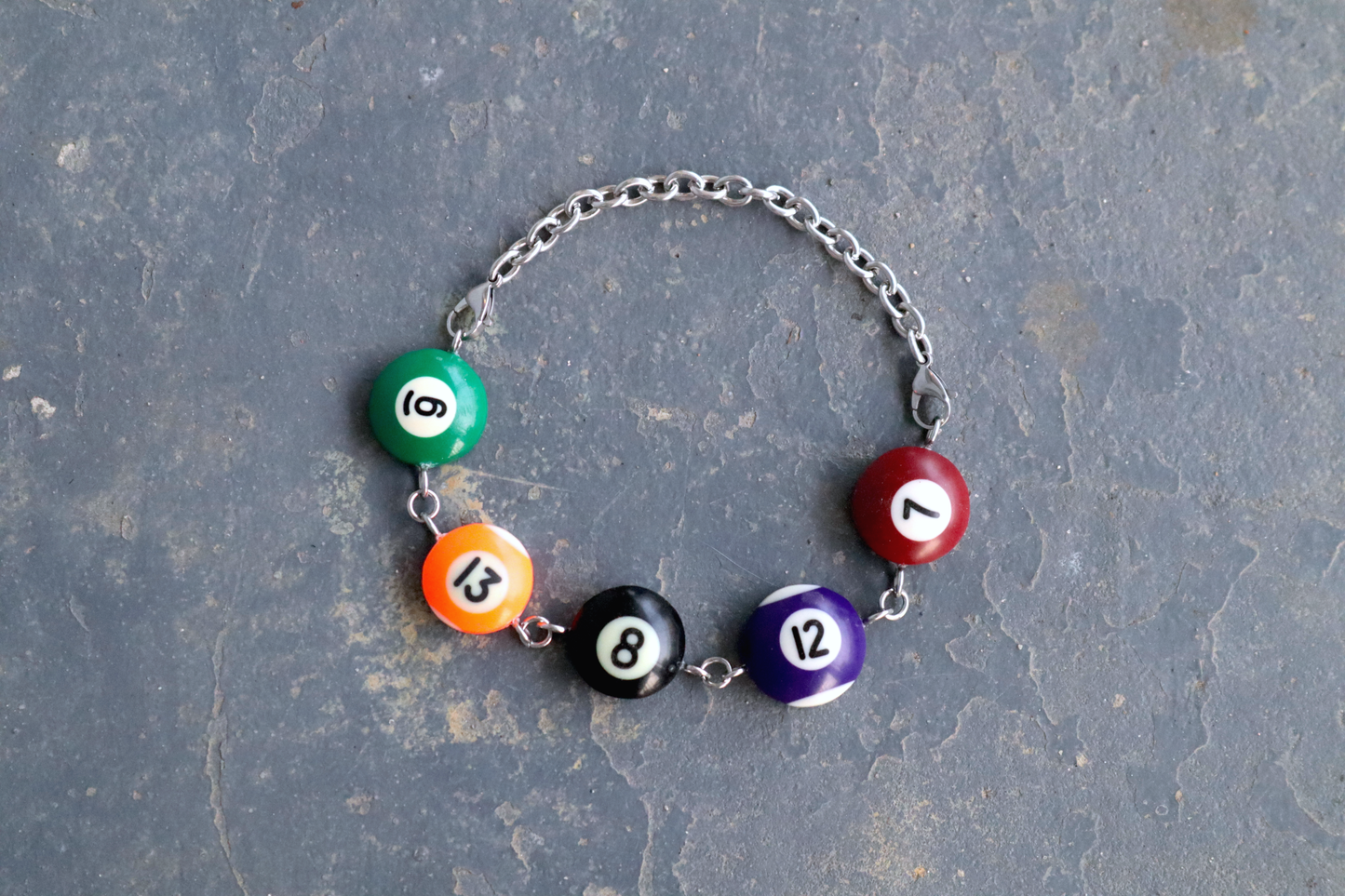 A choker made of pool ball charms numbered 6, 13, 8, 12 and 7 laid against a blue/gray surface. The choker has a steel chain and hardware to connect the charms.