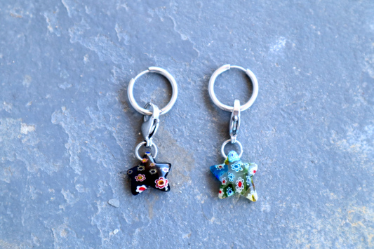 2 steel hoop earrings with 2 star-shaped pendants attached to them, resting against a blue surface.