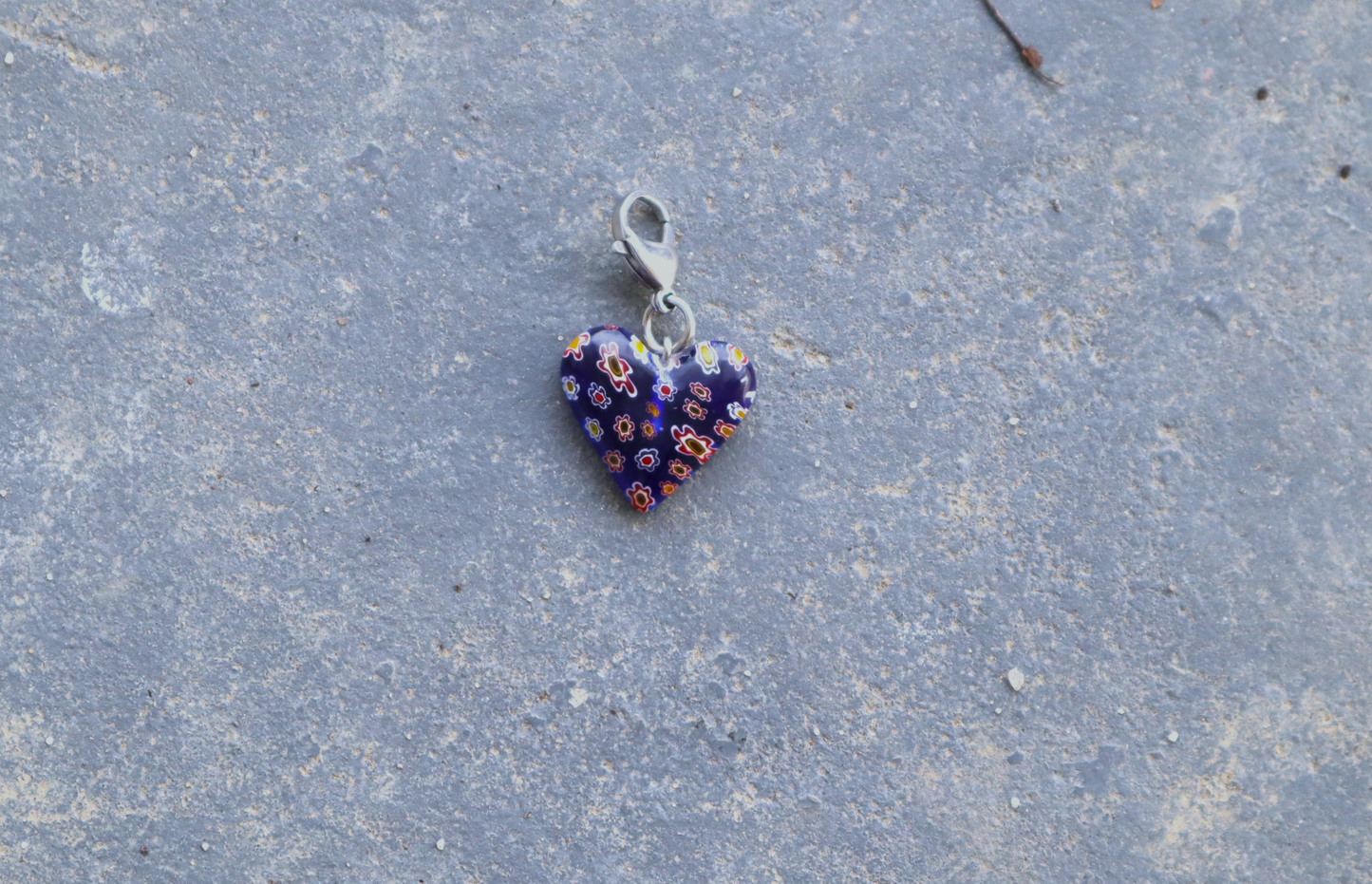 A heart-shaped blue pendant with white, red and yellow flower patterns. A silver clasp is attached to the top of the pendant, resting on a gray surface.