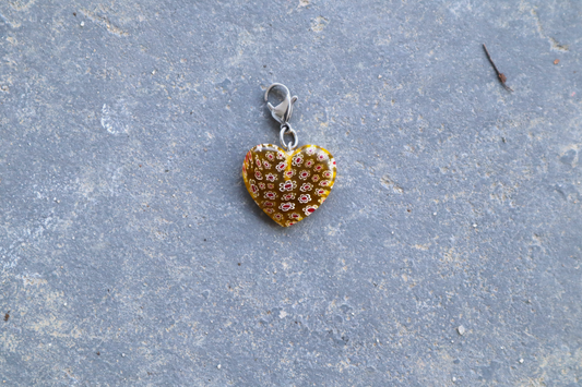 A heart-shaped yellow pendant with white and red flower patterns. A silver clasp is attached to the top of the pendant, resting on a gray surface.