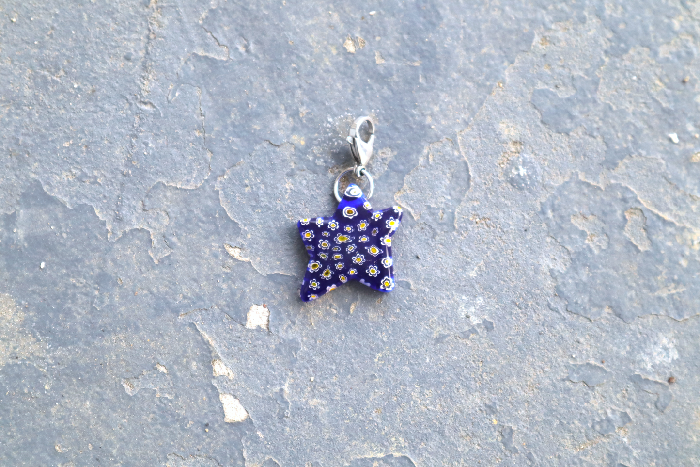 Star-shaped blue pendant with white and yellow flower patterns. A silver clasp is attached to the top of the pendant, resting on a gray surface.
