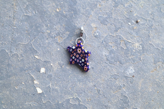 Star-shaped blue pendant with white, yellow and red flower patterns. A silver clasp is attached to the top of the pendant, resting on a gray surface.